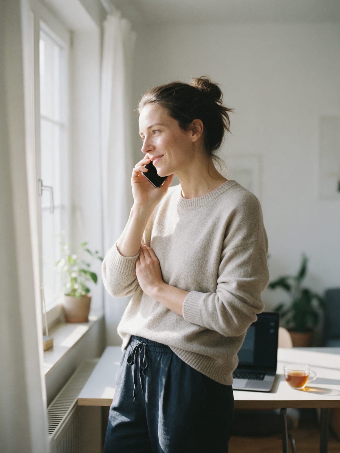 Eine Frau steht am Fenster ihrer Wohnung und telefoniert mit dem Smartphone.